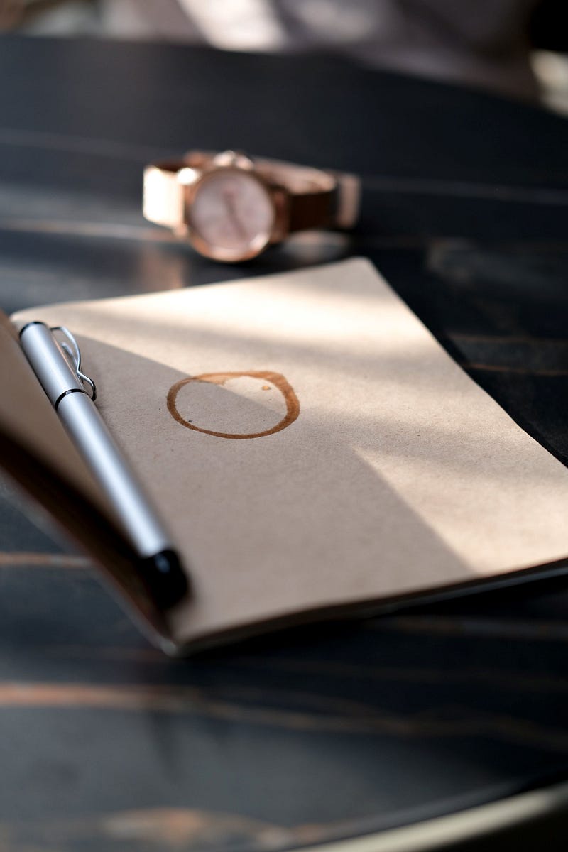 A pen rests atop an open notebook that has a coffee stain on its otherwise blank page.