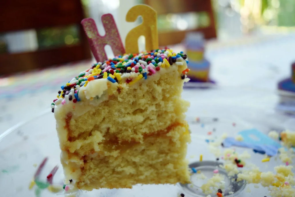 A close-up of a sprinkle-covered slice of birthday cake with the candle letters "Ha" on top, sitting on a crumb-covered plate.
